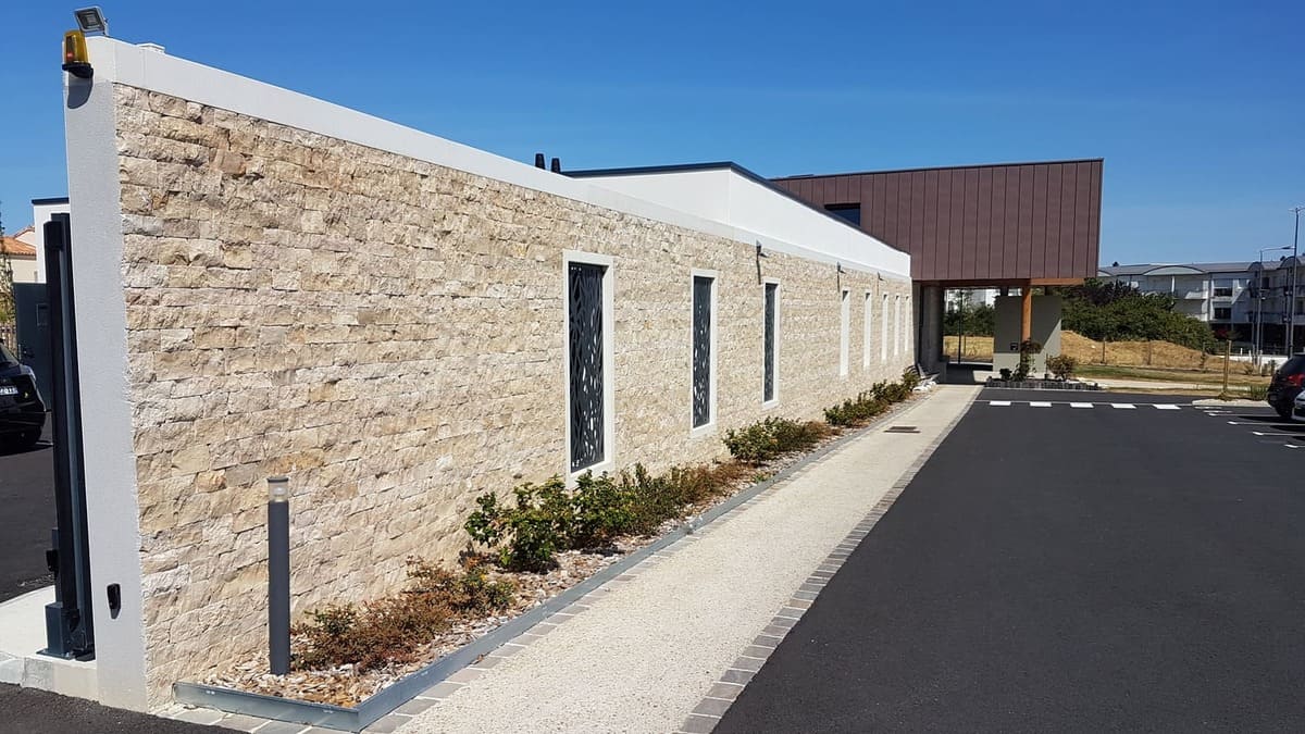 Façade extérieure en parement de moellons calcaires beiges à deux chants sciés, bâtiment neuf de réhabilitation psycho-sociale, avec claustra métalliques gris et massifs paysagers